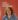 A headshot of a woman with shoulder length brown hair and hoop earrings with her glasses on her head and a blue dress on, in front of an orange poster which reads Leeds Climate Commission in white letters